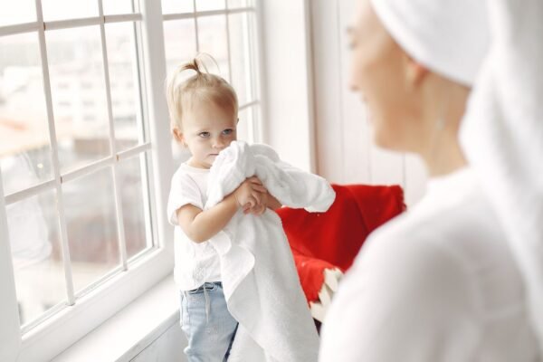 pexels-photo-5523115-5523115 Adorable toddler holding towels by a window with mother nearby, creating a warm family moment.