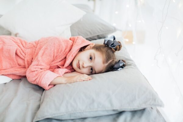 pexels-photo-6202206-6202206 A young girl in a cozy pink robe lying comfortably on a bed with soft pillows.